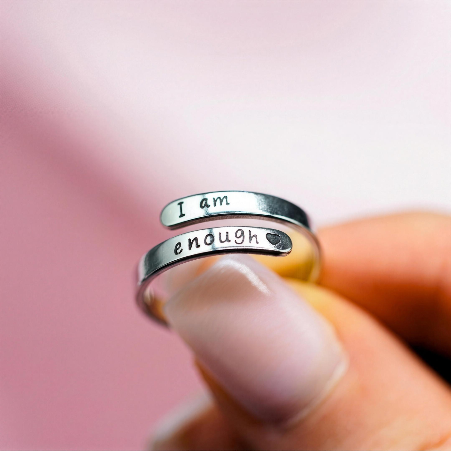I Am Enough Ring zwischen den Fingern gehalten, Gravur sichtbar, rosa Hintergrund